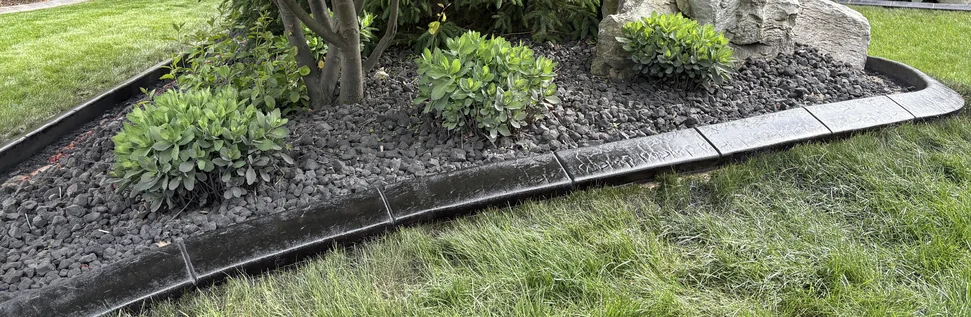Close-up of a matte black stamped concrete curb along a lava-rock foundation bed with dwarf shrubs.