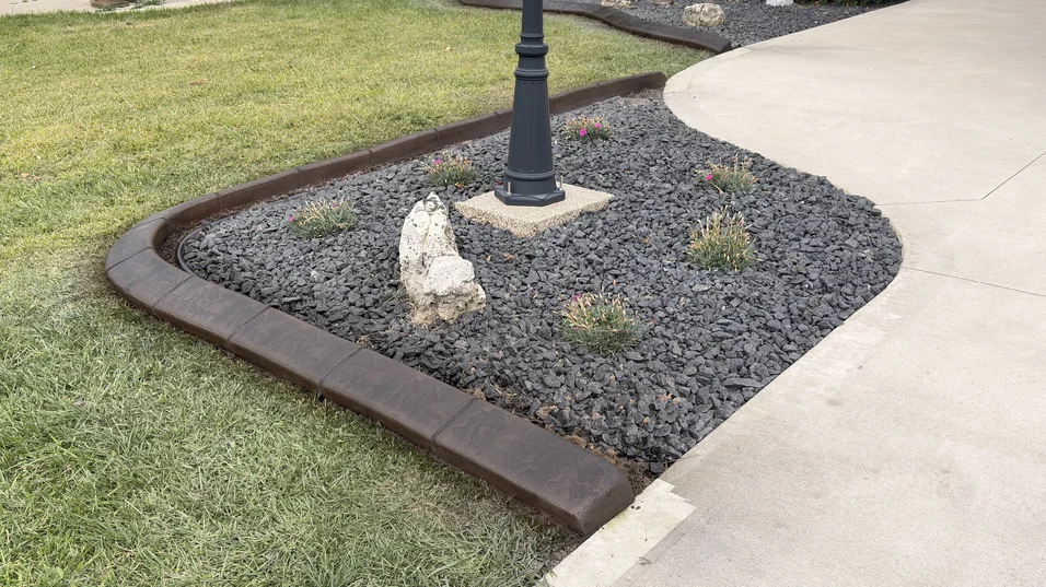 Warm brown stamped concrete curb around a black lava-rock bed featuring a decorative lamp post and accent boulder.