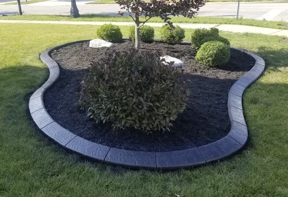 Charcoal stamped concrete curbing around a circular mulch island with ornamental shrubs on a front lawn.